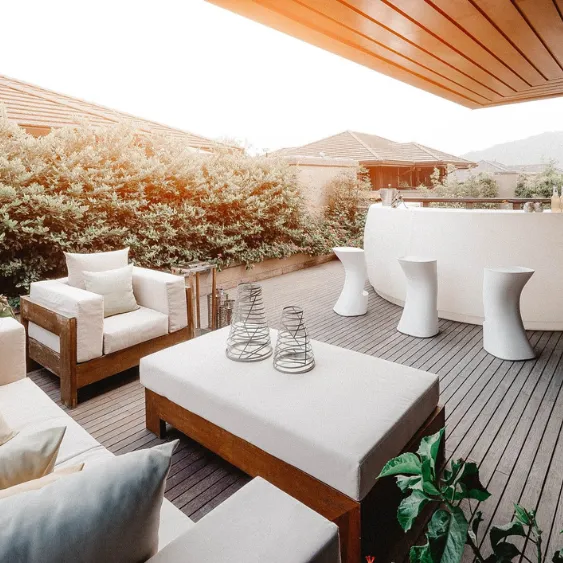 Patio moderne avec des meubles en bois et des coussins blancs, entouré de verdure, offrant un espace extérieur élégant et confortable.