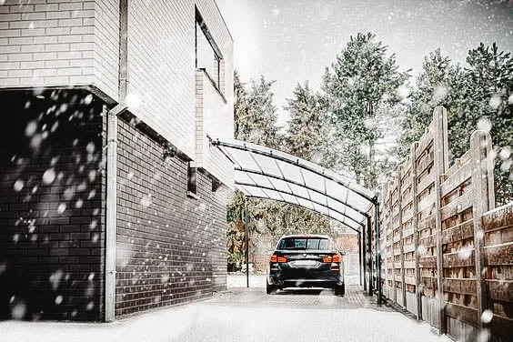 Voiture moderne garée sous un abri d’auto permanent avec un toit transparent, entourée de neige et d’arbres.