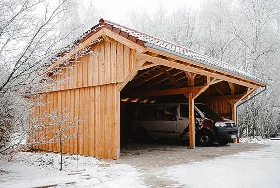 Abri d’auto en bois avec toit en pente, protégeant une fourgonnette sous un paysage hivernal enneigé.