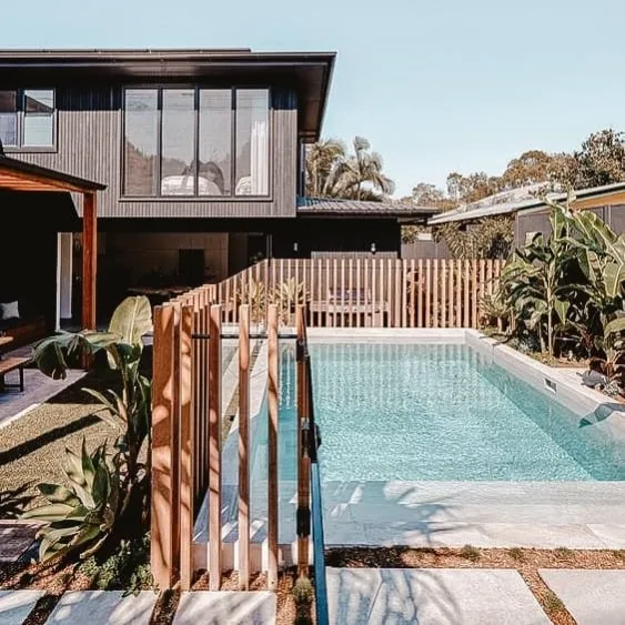 Modern pool area with a sleek wooden fence, surrounded by greenery and a contemporary home in the background.