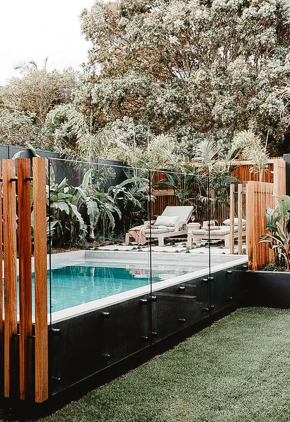 Modern pool area with glass fencing and wooden posts, surrounded by lush greenery and lounge chairs.