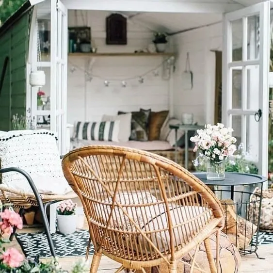 Cozy garden shed with open doors, featuring wicker chairs, a small table, and a beautifully decorated interior with cushions and plants.