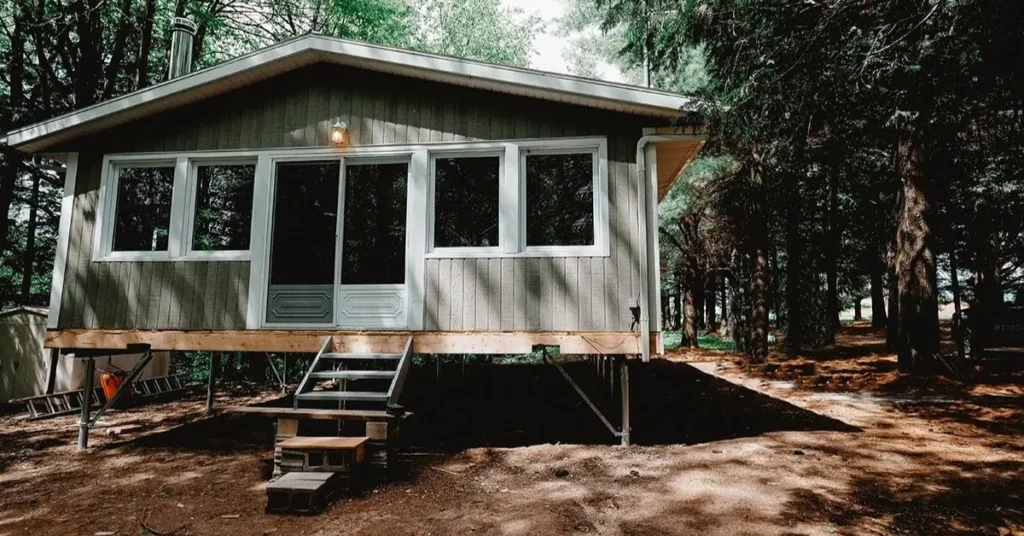 Small elevated cabin in a wooded area, supported by screw piles, with stairs leading to the entrance.