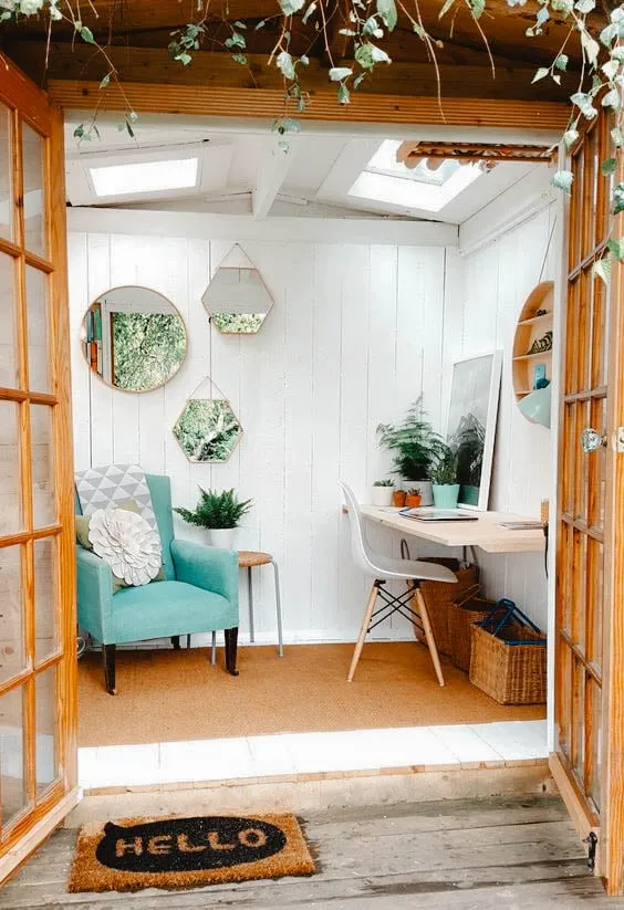 She Shed confortable avec un fauteuil turquoise, un petit bureau et des miroirs décoratifs sur des murs blancs, éclairée naturellement par des puits de lumière.