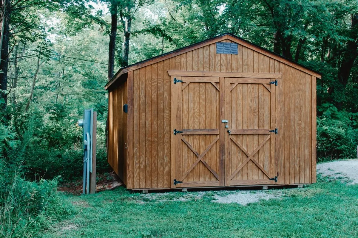 Cabanon de jardin en bois avec de grandes portes doubles, situé au milieu d'une forêt verdoyante.