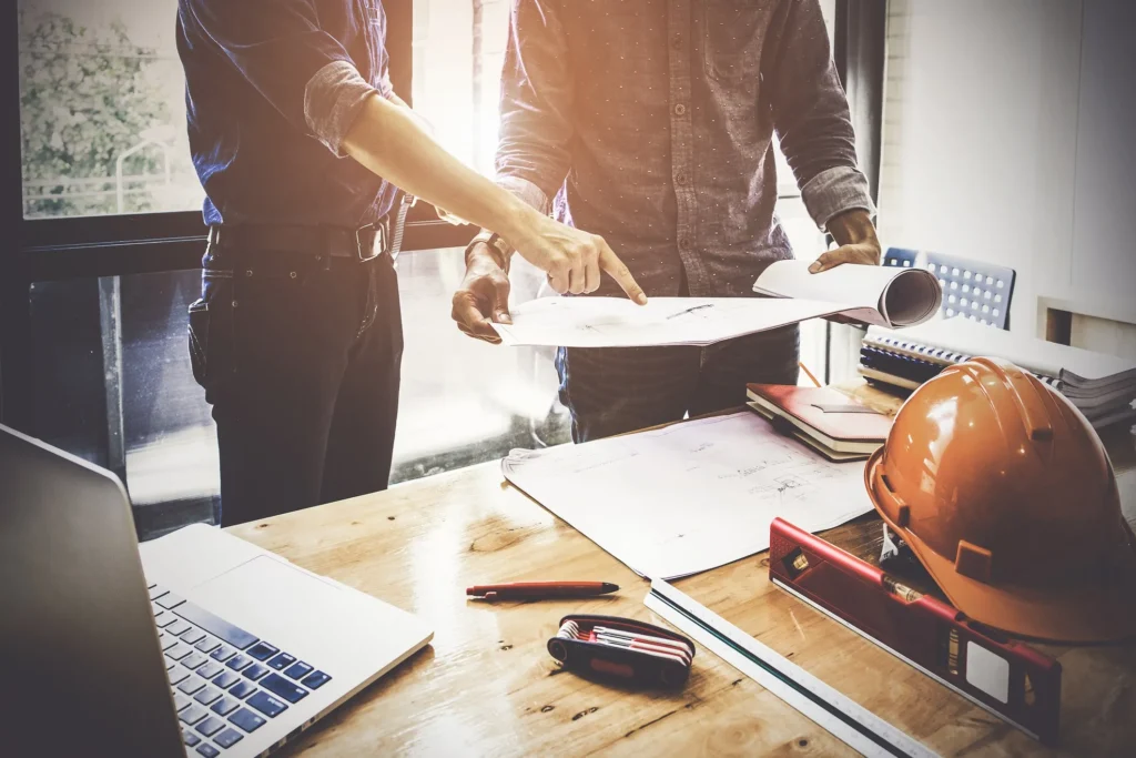 Deux professionnels examinant des plans de construction sur une table, avec un ordinateur portable et un casque de sécurité à proximité, illustrant une planification efficace.