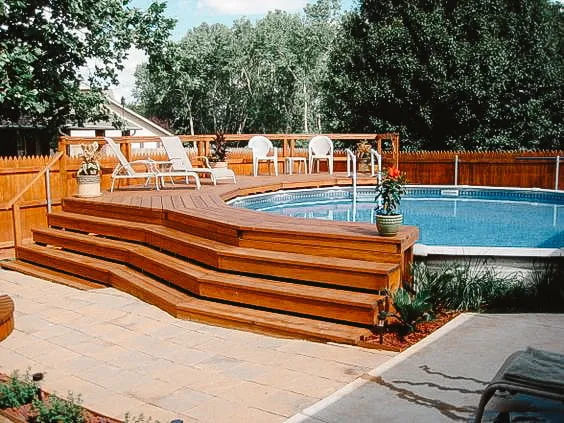 Terrasse en bois entourant une piscine hors-sol avec des marches à plusieurs niveaux et des chaises longues, située dans un jardin privé avec de la verdure.