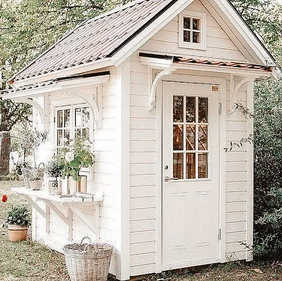 Petit cabanon blanc avec une porte vitrée et un toit à pignon, entouré de verdure et décoré de plantes en pots.