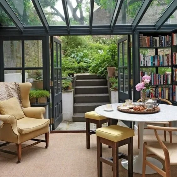 Elegant sunroom with a glass roof, bookshelf, and cozy furniture opening onto a lush garden.