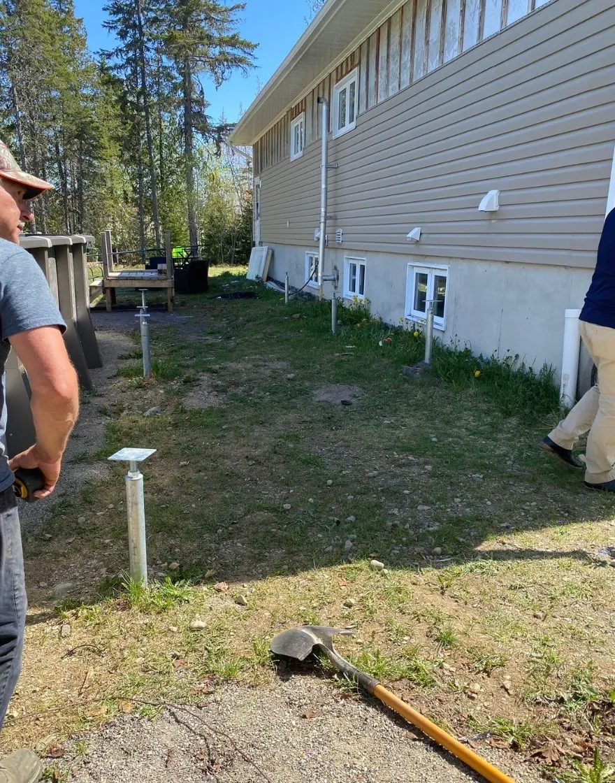 Installation of screw piles next to a house in preparation for building a stable and secure pool deck, with two workers assessing the area.