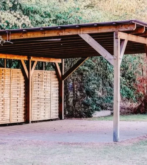 Carport en bois avec un toit solide et des panneaux de confidentialité, situé dans un jardin verdoyant et boisé.