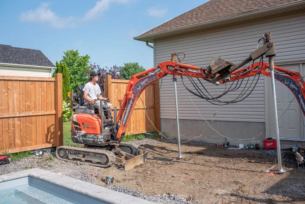 Travailleur utilisant un excavateur pour installer des pieux vissés dans une cour résidentielle, illustrant le processus de détermination de la profondeur idéale pour des fondations solides.