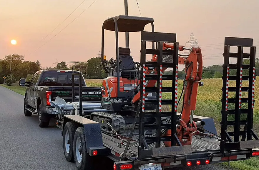 Camion Postech tirant une remorque avec une mini-pelle utilisée pour l'installation de pieux vissés au coucher du soleil.