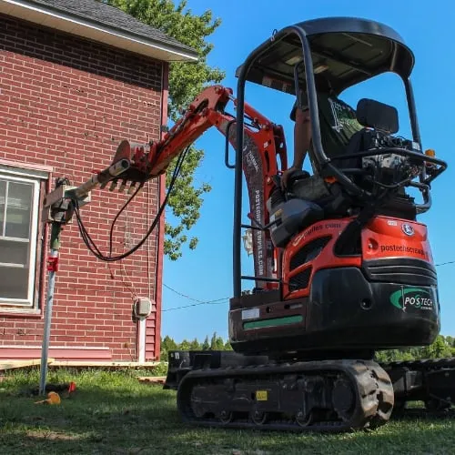 Mini-pelle orange installant un pieu vissé près d'une maison en briques rouges.