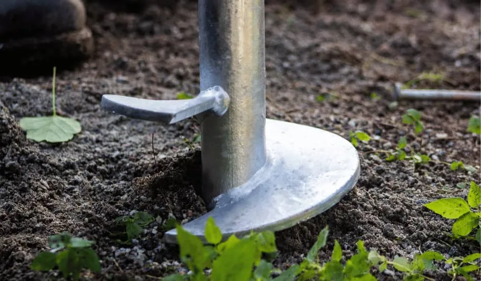 Gros plan d'un pieu vissé en métal partiellement enfoncé dans le sol avec des plantes vertes autour.