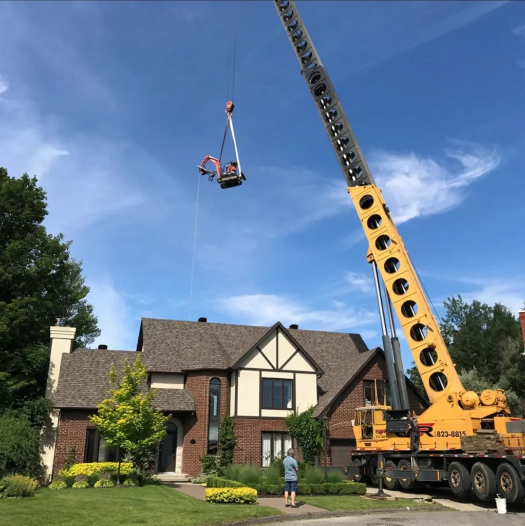 Excavator being lifted by a large crane over a residential house with a man observing from the front yard on a clear day.