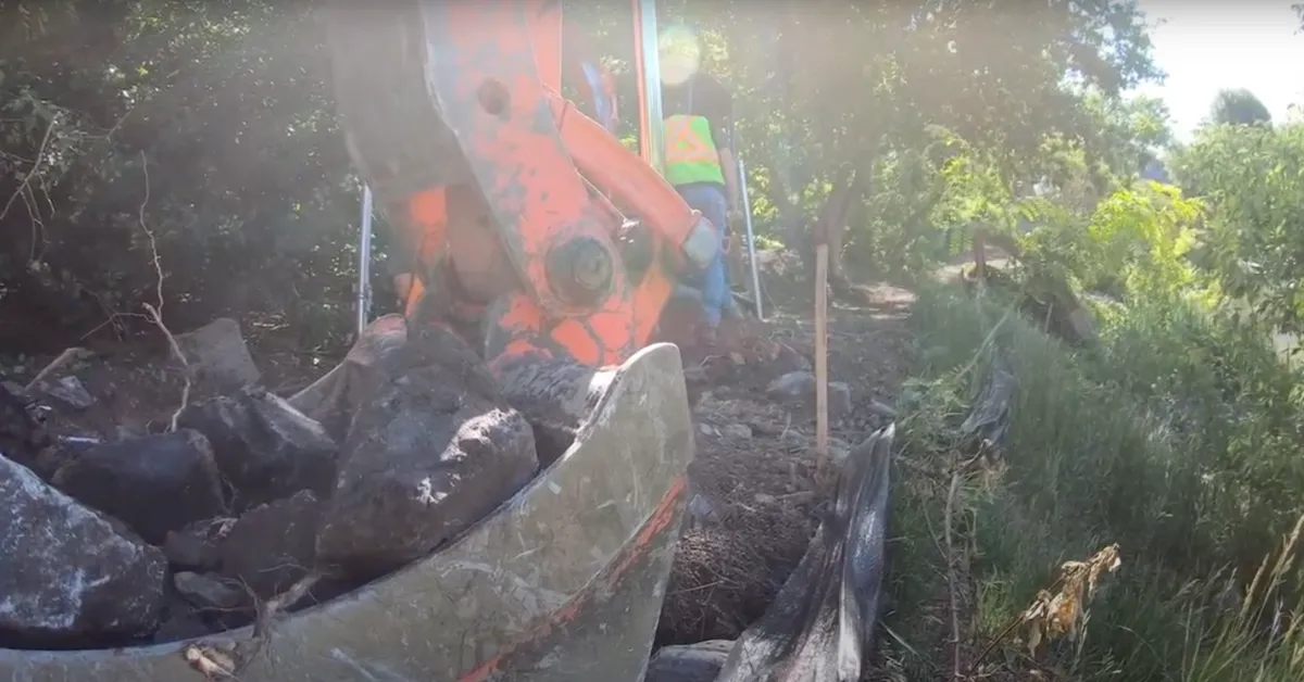 Godet d'excavatrice rempli de grosses roches, travaillant sur un terrain rocailleux avec des ouvriers en tenue de sécurité à proximité dans une zone boisée.